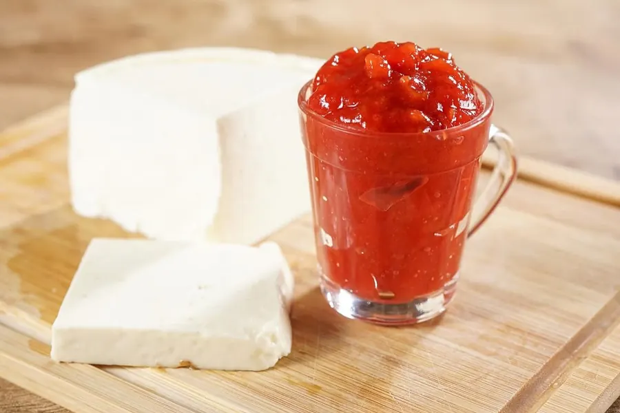 Queijo minas em fatias grossas na tábua de madeira, ao lado de um copo de goiabada cascão em pedaços.