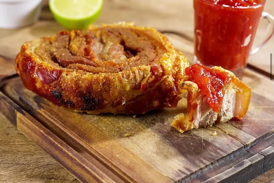 Torresmo de rolo gourmet na tábua de madeira, com uma fatia aberta coberta de geleia de pimenta e limão ao fundo.
